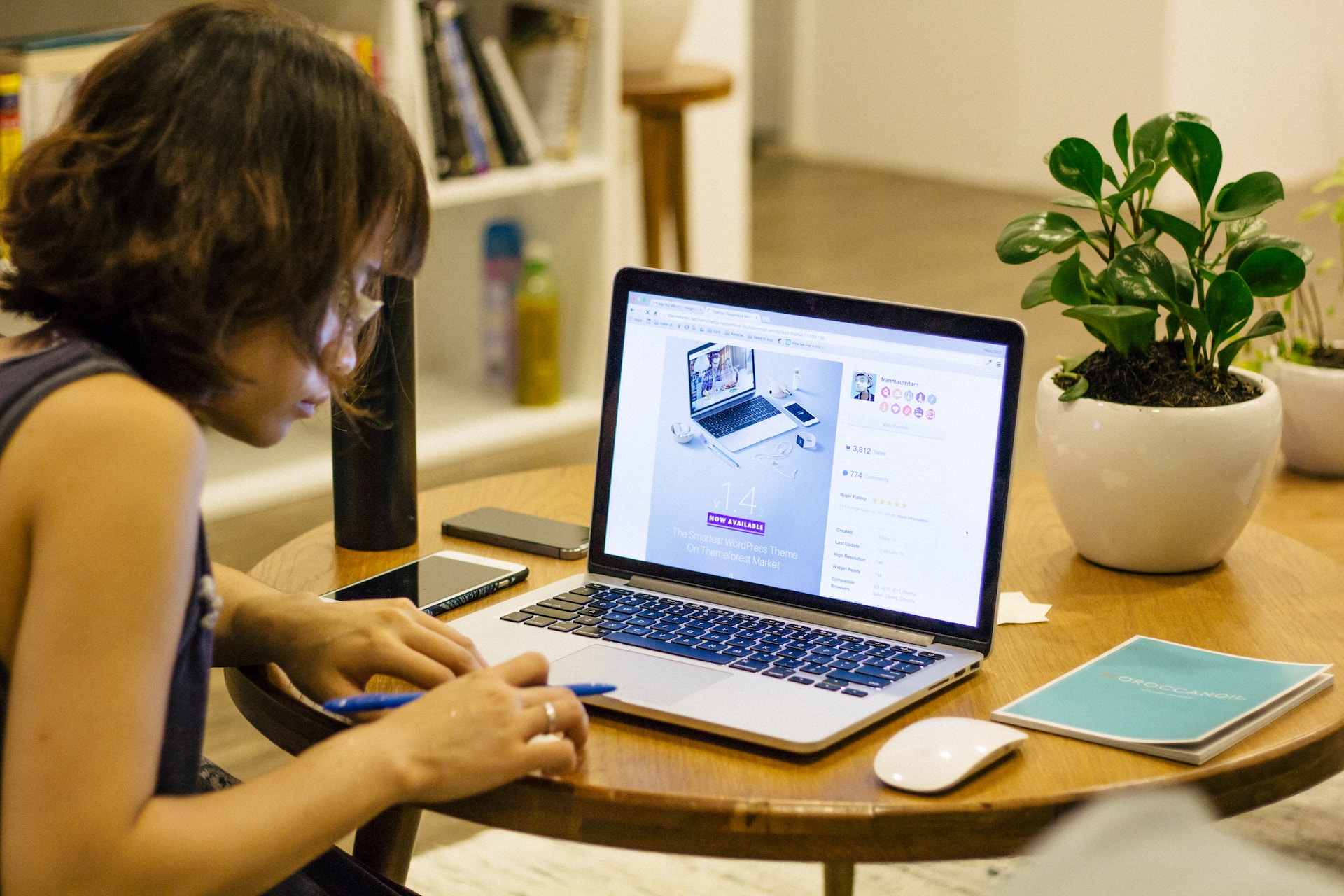 Asian woman with glasses using a laptop on a light wood table with a plant.